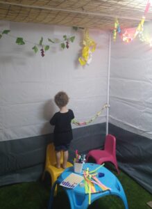 ילד עומד על שרפרף ומקשט את דופן הסוכה, לצדו שולחן עם טושים וחומרי יצירה