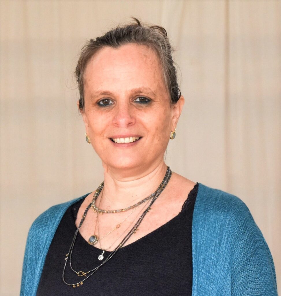 אישה חיוכת פנים למצלמה, לבושה בחולצה שחורה וסוודר כחול, עם תכשיטים