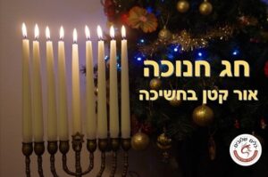 חנוכייה עם נרות לבנים דולקים על רקע עיטורים מוארים, לצד הכיתוב חג חנוכה – אור קטן בחשיכה