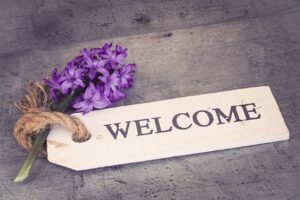 שלט עץ לבן עם הכיתוב WELCOME מונח על משטח אפור, לצדו זר פרחי יקינטון סגולים