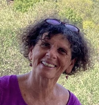 דיוקנה של עפרה גורן מחייכת בחוץ, משקפיים מורמים על ראשה וחולצה סגולה
